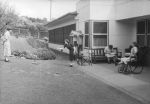 Long shot of four people outside a building two people are playing a game of quoits, one person is seated on a wooden bench reading a book, one person is in wheelchair with calipers on legs. Ring Toss on the Lawn, Circa 1955