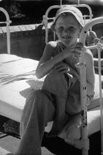 Medium shot of a two children outside one child in the foreground is seated on a bed with one hand holding the end bed rail wearing a hat, the other child is in the background laying supine on a bed, a small brick wall is behind the beds. In the Sunshine, Circa 1955