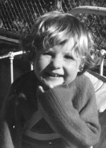 Medium shot of a child outside seated on a bed with braces fitted to neck and chest posing for the camera, a wire fence is in the background. Portrait of a Child, Circa 1955