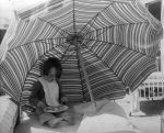 Long shot of a child wearing a bib and eating from a plate of food while sitting under a striped beach umbrella. Dinner Under the Umbrella, Circa 1955