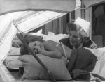 Medium shot of a child lying face down on a bed. A nurse is sitting behind and they are both shaded by a striped beach umbrella. Under the Umbrella, Circa 1955