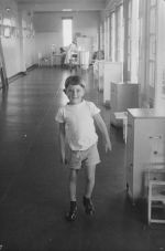 Long shot of a child walking down a long room. There is a bed at the very end and small chests of drawers along the side next to the windows. In the Ward, Circa 1955