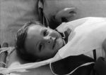 Close shot of a child lying down facing the camera. There is some sort of top tied to the bed head and covering the chest. Portrait of a Child, Circa 1955