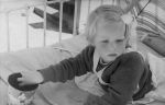 Close shot of a young child lying on a bed out in the sun with a beach umbrella. There is a small toy pail on the bed. Portrait of a Child, Circa 1955