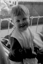 Medium shot of a young child smiling at the camera sitting on a bed wearing a large bib. Portrait of a Child, Circa 1955