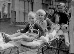 Long shot of five children sitting one behind the other on a long bed with large spoked wheels. The photograph is taken outside and shows just the side of a nurse standing next to the bed. Three of the children have their leg in a cast. Posing on a Trolley, Circa 1955