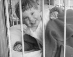 Close shot through the bars of a cot of a young child holding a blanket and smiling at the camera. Portrait of a Child, Circa 1955