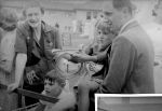 Medium shot of two children . One is lying face down and the other is being held by a man. The child is holding a looped rope. There is also a woman standing on the other side of the children. The photograph is taken outside and there are other people in the background. Ring Toss, Circa 1955