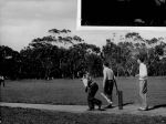 A long shot of three men playing a game of cricket. The photograph shows the bowler, batter and umpire at one end of the pitch. Playing Cricket, Circa 1955