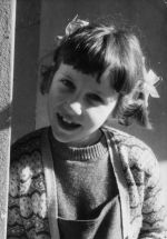 Close up shot of a young child smiling for the camera. The child has ribbons in their hair and is wearing a fair isle cardigan. Portrait of a Child, Circa 1955