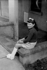 A medium shot of a young patient sitting outside on a brick porch. The left leg is in a cast. Portrait of a Child, Circa 1955