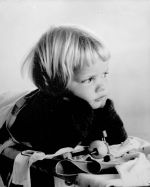A close up shot of a young child lying on their stomach with their head propped up by their arms. They appear to be lying on a stretcher. Portrait of a Child, Circa 1955