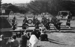 A long shot of a band marching across grass. Band members are wearing kilts, holding musical instruments. There are people sitting in the foreground (backs to the camera) watching the band, and there are swing sets and a see-saw in the background. Watching the Pipe Band, Circa 1955