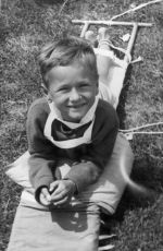 A medium shot of a young child lying on their stomach on a stretcher on the ground. They are attached to the stretcher by ties around their calves. Outside on the Lawn, Circa 1955