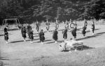 A long shot taken outside of a band. Band members are in a circle, playing musical instruments. They are being watched by two children lying on their stomachs in beds, with a man squatting down beside the left hand side bed. Hawthorn Pipe Band, Circa 1955