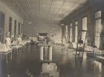 Long shot of a nightingale style ward. Running down the centre of the image is a table and numerous vases of flowers. The left and right walls are lined with patient beds, some beds are occupied with children. Several Nurses are standing next to beds. Ward Scene, 1923-1924