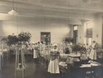 Wide shot of a Nightingale style ward with young children and babies. Nurses holding and attending children. Large vases of flowers/foliage throughout can be seen. Ward Scene, 1923-1924