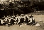 Long shot of adults sitting down in a group on a grassed area with trees and shrubs in the background. School Group on Lawn, 1923