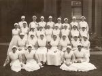 Long shot of a group of staff members, including four sitting on ground, seven sitting on chairs in the first row, nine standing up in third row and eight standing in the back row. Staff members are in uniform. Photograph was taken outside in front of a building. Nursing Staff Portrait, Circa 1920