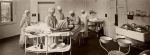 Long shot of interior of hospital room. Bed is positioned in the middle with patient lying on the bed, staff are positioned around the bed. The room consists of medical equipment and technology of that era. Operation in Progress, Circa 1920