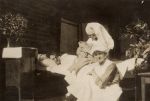 Long shot of three adults, one adult laying in the bed and two staff members. One staff member is holding an infant. Candid Ward Scene, 1922