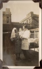 Long shop of two staff members standing one staff member is holding a young child. A cot and buildings are present in the background. Medical Staff and Patient, Circa 1941