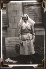 Long shot of a staff member in uniform standing against a brick wall, plaque with writing and garden bed. Sister Outside the Hospital, Circa 1945