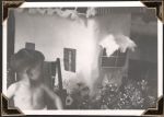 Close up of a child sitting to the left of the frame. He looks behind him at a paper-makeshift cubby house and an array of flowers on the ground. Play Time, Circa 1945
