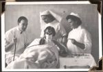 Medium shot of three hospital staff in uniform; one male and two female. They stand around a patient who sits up in their bed. The age of the patient is unclear, either teenage or adult. They all face the camera front on. Surgery, Circa 1945