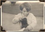 Medium shot, taken outside, of a young child holding two objects and looking down. There are a path, grass and shrub/tree behind them. Outdoors, Circa 1945