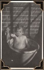 Medium shot of young child sitting in large bucket against a brick wall, holding up a washer in their right hand. Infant in Tub, Circa 1945