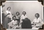 Medium shot of five women. One standing on the left of the photograph and four sitting to her left. Three of these look to be in nurse's uniforms. Foreground shows what may be the back of another woman's head. Farewell, Circa 1945