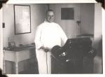 Medium shot of man in a gown posing in front of machine with controls in his right hand. Desk visible behind him with a framed print on the wall above desk. Door visible over his left shoulder. X-Ray, Circa 1945