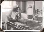 Medium shot of a staff member leaning over the head of young child patient lying on bed with arms raised above head. Sink visible on right side of photograph. Window visible on left side of photograph. Chest X-Ray, Circa 1945