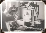 Medium shot of two staff members with young child lying on bed with arms above head. One staff member is holding child's arms. Another staff member is guiding machine placed above child's head. Chest X-Ray, Circa 1945