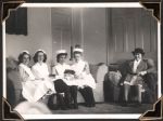 Medium shot of four staff members, in uniforms, sitting together on a couch and a woman wearing a coat and hat sitting on a lounge chair. Doors are visible in the background. Nurses Lounge, Circa 1945