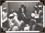 Medium shot of seven staff members, some holding a cup and saucer, seated around a seated woman wearing a hat and coat, holding a cup and saucer. Double wooden doors are in the background. Nurses Lounge, Circa 1945