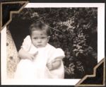 Close up shot of an infant wearing a white gown, eyes open, taken outside in front of a bush. An Infant, Circa 1945