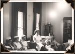 Medium shot of six seated staff members, taken in a room with tall windows in the background on the left hand side, and a bookshelf with glass doors is on the right hand side. There is a seventh staff member on the right hand side, sitting on a chair but is only partially visible. Nurses Lounge, Circa 1945