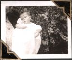 Close up shot of an infant wearing a white gown, head leaning towards right shoulder, taken outside in front of a bush. An Infant, Circa 1945