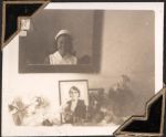 Medium shot of a staff member's face in a mirror on a wall. Below the mirror are a number of artefacts including framed photographs and flowers. Self Portrait, Circa 1945