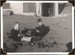 Long shot of two children sitting on the ground cleaning shoes. The façade of a building can be seen with a door and gardening tools leant against it. Shoe Cleaning, Circa 1945