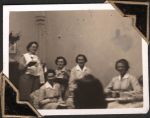 Long shot of six female hospital staff gathered in a room. Five are sitting, one of which faces away from the camera, the back of her head is close to the camera. The remaining five all face the camera, one of which stands. Nurses Gathering, Circa 1945