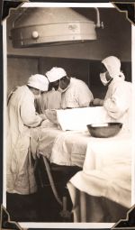 Long shot of four medical staff dressed in gowns, masks and hair caps, around a medical bed. Three of the staff are easily seen while one standings behind them, out of full view. They are all attending to something on the bed which cannot be seen. Operation in Progress, Circa 1945