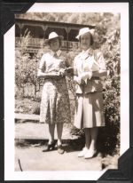 Long shot of two women in casual dress and hats standing side by side outside a two-story building with a porch and verandah on the upper level. Trees can also be seen around the ladies. Two Females in the Sunshine, Circa 1945