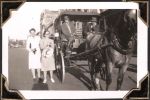 Long shot of a horse-drawn carriage with two men sitting in the front and two women in dresses standing next to it. The view of a street can be seen in the background. Horse Drawn Carriage, Circa 1945