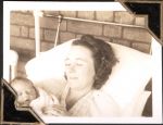 Close up of a lady lying in a hospital bed holding a new born baby. The bed is a white metal frame and it is against a brick wall. Women's Hospital, Circa 1946
