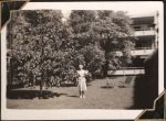 Long shot of a woman standing outside a hospital building amongst trees. She wears a skirt and blazer and is holding an unknown item. Women's Hospital, Circa 1946