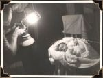 Medium shot of a newborn infant in a cot with blankets covering them. They are to the right of the photograph. On the left there is an adult holding a light. The infant gazes at the light. Women's Hospital, Circa 1946