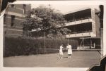 Long shot of two women standing on a tennis court, one on either side of the net as they shake hands. They wear white tennis attire. In the background are surrounding buildings and trees. Women's Hospital, Circa 1946
